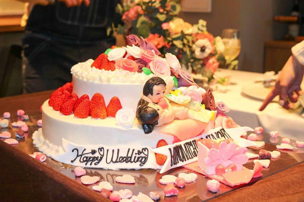 ウェディングケーキの製作事例。2段の苺のウェディングケーキ。前にリボンを飾りhappy weddingと新郎新婦のお名前。ボックスの中に沢山のハート型を入れて…ケーキの上には新郎が新婦を抱っこしているマジパン細工のお人形。ケーキの周りにはハートがいっぱい。そんなウェディングケーキの写真。